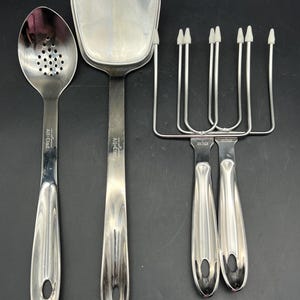 May include: A set of stainless steel kitchen utensils, including a slotted spoon, a spatula, and a carving fork. The utensils have long, curved handles and are designed for cooking and serving food. The brand name "All-Clad" is visible on the spoon and spatula.