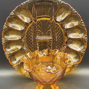 May include: Amber-colored glass serving dish with a round, scalloped edge and multiple compartments. A small, ruffled bowl with three feet is attached to the front. The dish has a textured pattern throughout.