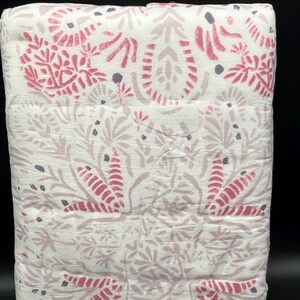 May include: A folded white quilt with a repeating floral pattern in shades of pink and gray. The design features stylized leaves and floral elements, creating a soft, textured appearance. The quilt is neatly stacked, showcasing the detailed print.