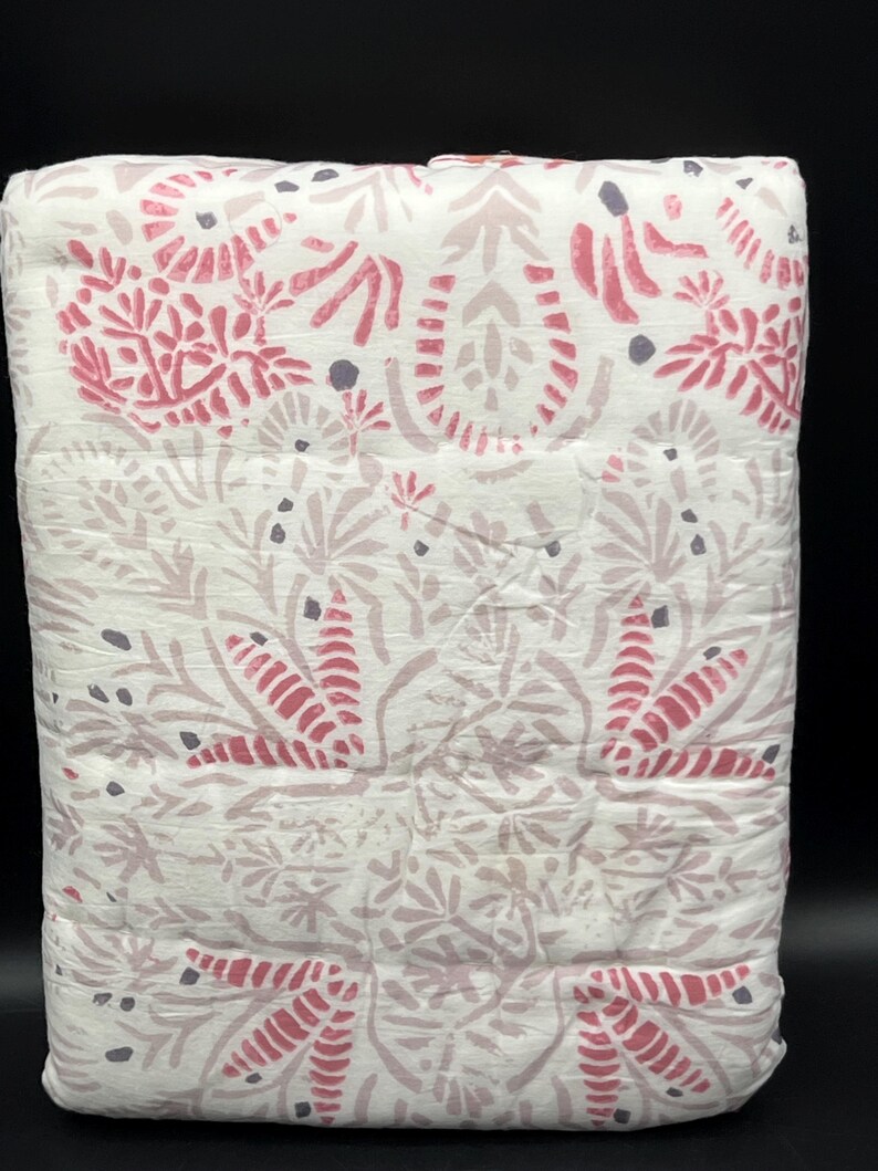 May include: A folded white quilt with a repeating floral pattern in shades of pink and gray. The design features stylized leaves and floral elements, creating a soft, textured appearance. The quilt is neatly stacked, showcasing the detailed print.