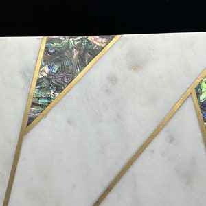 Op de afbeelding: Close-up van een wit marmeren oppervlak met ingelegd abalonschelp en goudkleurige metalen accenten. Het ontwerp heeft geometrische vormen met iriserende schelpsegmenten en dunne metalen lijnen, wat een luxe uitstraling cre&euml;ert.