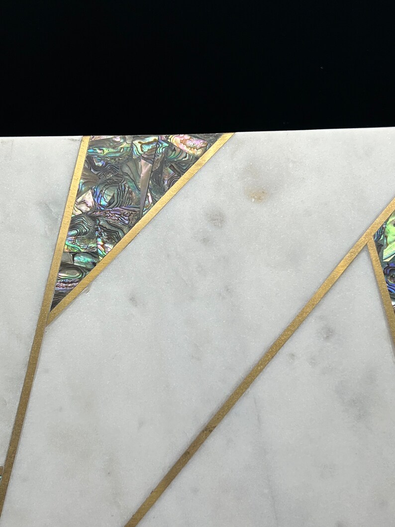 Op de afbeelding: Close-up van een wit marmeren oppervlak met ingelegd abalonschelp en goudkleurige metalen accenten. Het ontwerp heeft geometrische vormen met iriserende schelpsegmenten en dunne metalen lijnen, wat een luxe uitstraling cre&euml;ert.