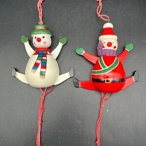 May include: Two wooden jumping jack ornaments featuring a snowman and Santa Claus. The snowman is white with a green hat and colorful scarf. Santa wears a red suit with a green trim and blue striped scarf. Both have red pull strings attached.