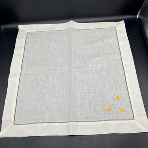 May include: White linen napkin with a yellow embroidered design of three pineapples.
