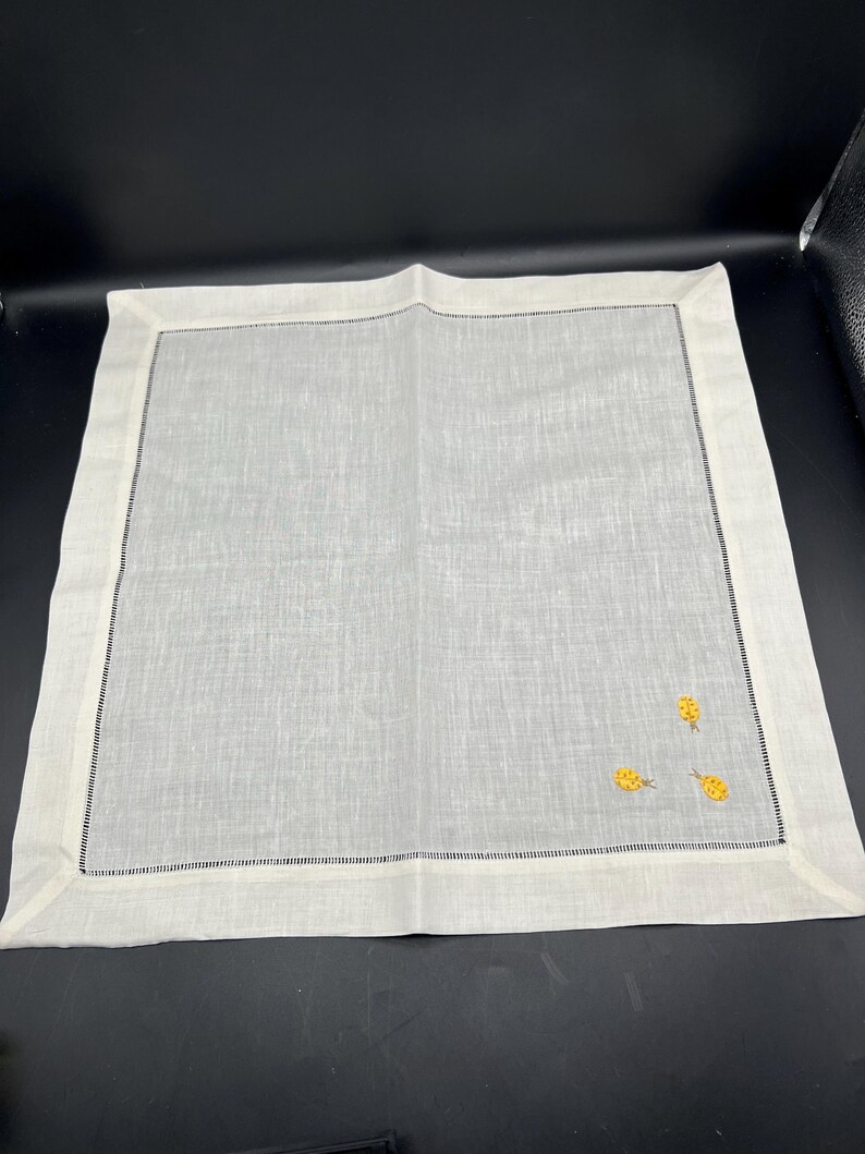 May include: White linen napkin with a yellow embroidered design of three pineapples.
