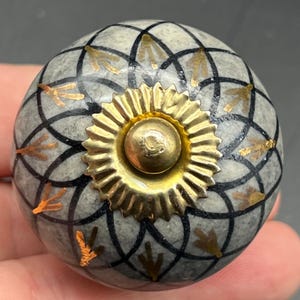 May include: A decorative cabinet knob with a gray and black geometric pattern and gold accents. The knob has a gold-colored center and is approximately 1.5 inches in diameter. The knob is round and has a textured surface.