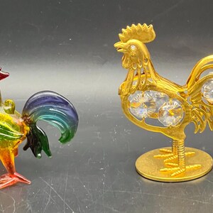 May include: A multicolored glass rooster figurine with a red comb and a blue-green tail, positioned next to a gold-toned metal rooster figurine featuring clear, faceted crystal accents embedded in its body and a circular base.