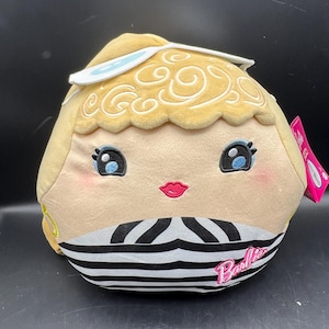 May include: A plush doll in the shape of a Barbie doll head. The doll has blonde hair, blue eyes, and a pink mouth. The bottom of the doll is black and white striped with the word "Barbie" printed on it.