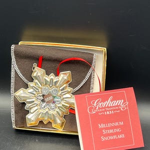 May include: A silver and gold snowflake ornament with a red ribbon, presented in a brown box with a gold trim. The ornament is labeled "2000". A red card reads "Gorham A Holiday Tradition Since 1831" and "Millennium Sterling Snowflake."