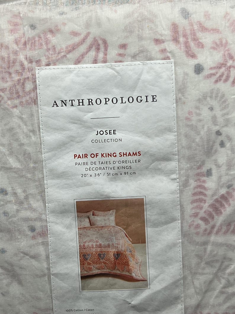 May include: A tag from Anthropologie for a pair of king shams. The tag reads "JOSEE COLLECTION" and "PAIR OF KING SHAMS". The shams measure 20" x 36" (51 cm x 91 cm). The background shows a floral pattern.
