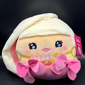 May include: A plush Barbie doll with a cream-colored bonnet and pink accents. The doll features a smiling face with blue eyes, a gold necklace, and two large pink bows. The Barbie logo is visible.