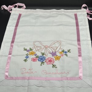 May include: White embroidered baby bib with a pink bow and floral design. The bib features pink ribbon trim and the embroidered text "Porta Biancheria". It has pink ribbon ties at the top.