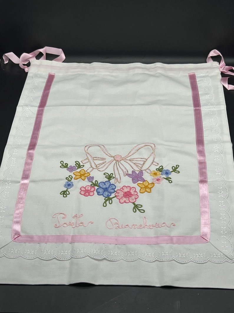 May include: White embroidered baby bib with a pink bow and floral design. The bib features pink ribbon trim and the embroidered text "Porta Biancheria". It has pink ribbon ties at the top.
