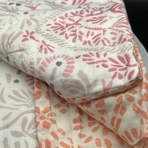 May include: A folded quilt with a floral pattern in shades of pink, peach, and lavender on a white background. The quilt has a quilted design and a soft, inviting appearance. The floral pattern is repeated throughout the quilt.