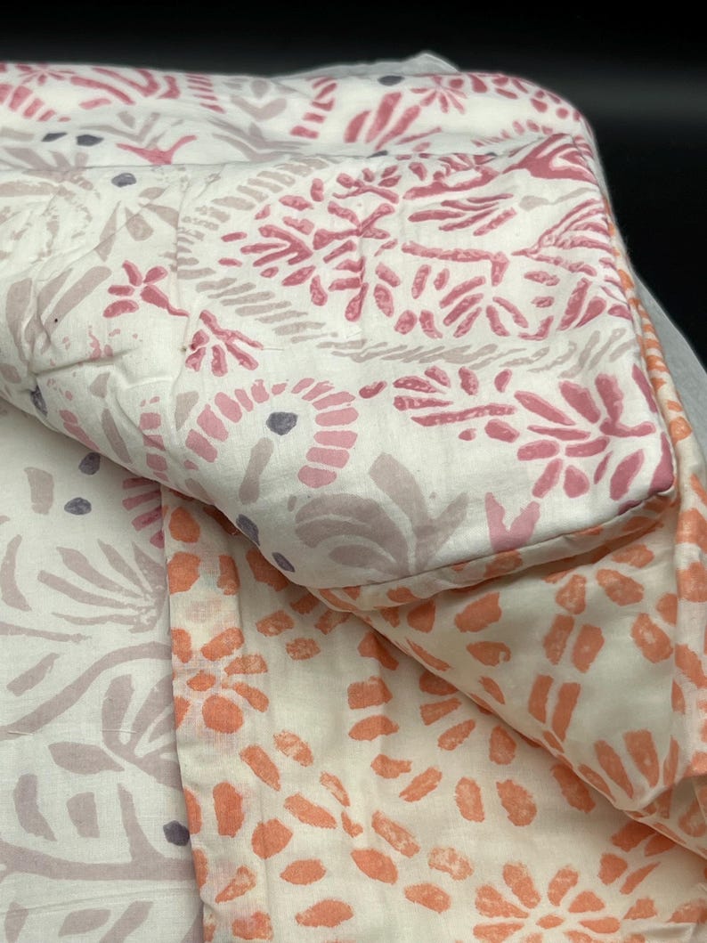 May include: A folded quilt with a floral pattern in shades of pink, peach, and lavender on a white background. The quilt has a quilted design and a soft, inviting appearance. The floral pattern is repeated throughout the quilt.