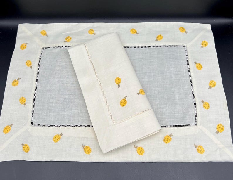 May include: A white linen table runner with a white linen napkin. Both are embroidered with yellow pineapple designs.