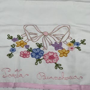 May include: White embroidered fabric featuring a pink bow and floral arrangement in shades of pink, blue, yellow, and purple. The text "Porta Biancheria" is embroidered in pink. A pink ribbon and a scalloped lace trim complete the design.