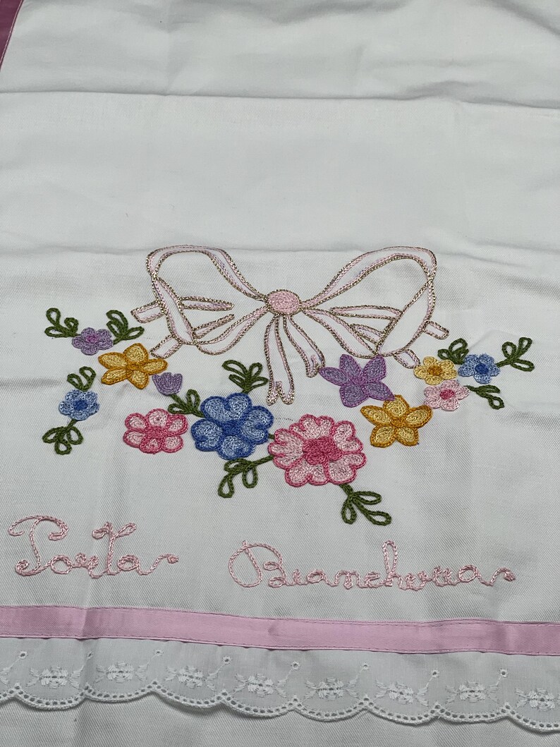 May include: White embroidered fabric featuring a pink bow and floral arrangement in shades of pink, blue, yellow, and purple. The text "Porta Biancheria" is embroidered in pink. A pink ribbon and a scalloped lace trim complete the design.