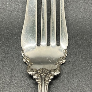 May include: A silver fork with an ornate handle and four tines. The fork has a decorative design on the handle and the back of the fork is marked with the number "92".