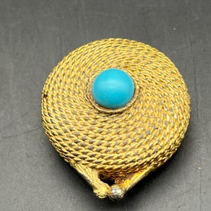 May include: A vintage gold-toned brooch with a woven rope design. The brooch features a round, bright turquoise stone at the center. The clasp is silver-toned. The brooch is likely from the mid-century era.