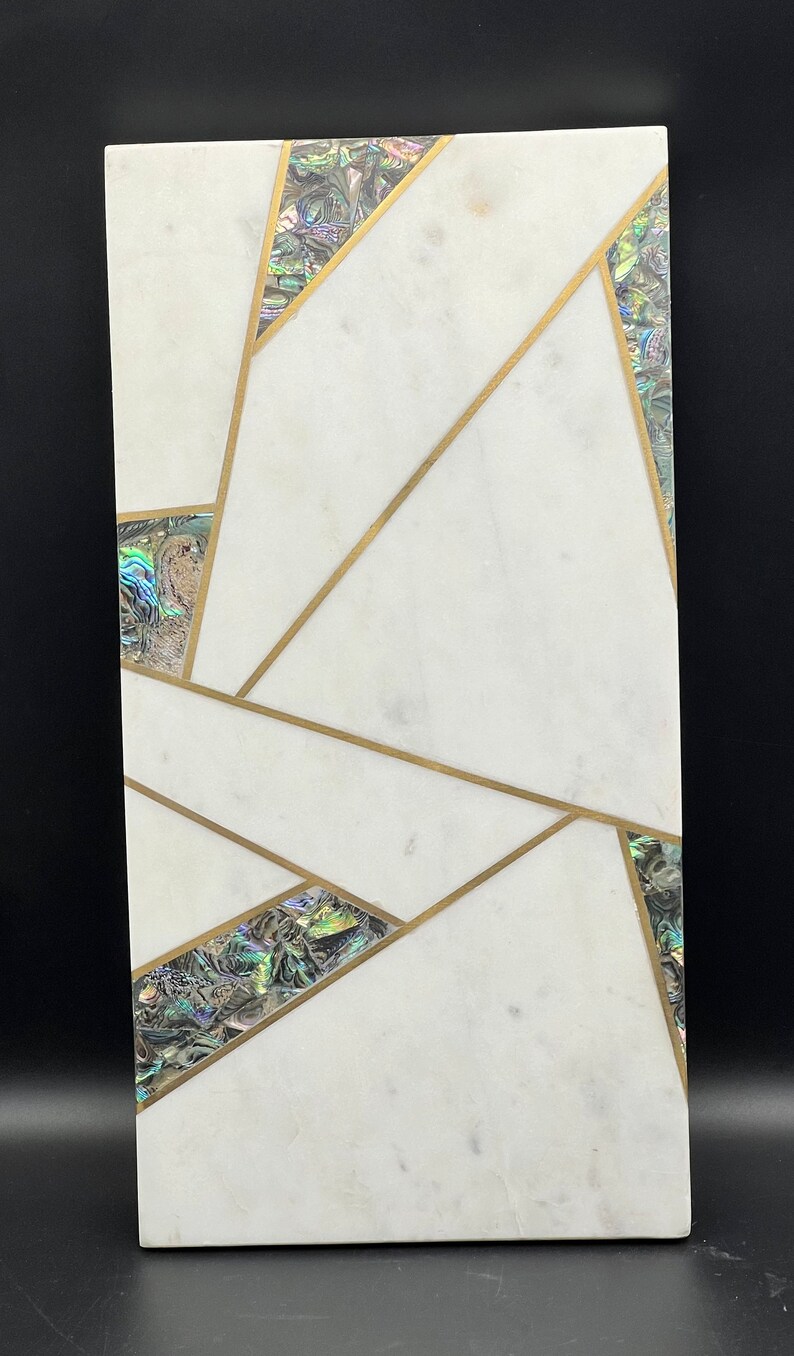 Op de afbeelding: Rechthoekig wit marmeren serveerplank met geometrische gouden inleg en iriserende abalone schelp accenten. Het bord is een decoratief stuk voor het serveren van eten of het uitstallen van items.