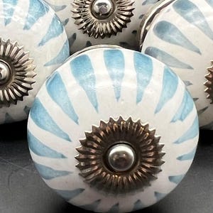 May include: Close-up of several white ceramic cabinet knobs with a blue striped pattern. Each knob has a decorative metal base and a silver-colored center. The knobs are likely for furniture or decorative use.
