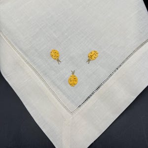 May include: A white linen napkin with a delicate embroidered design of three yellow pineapples.