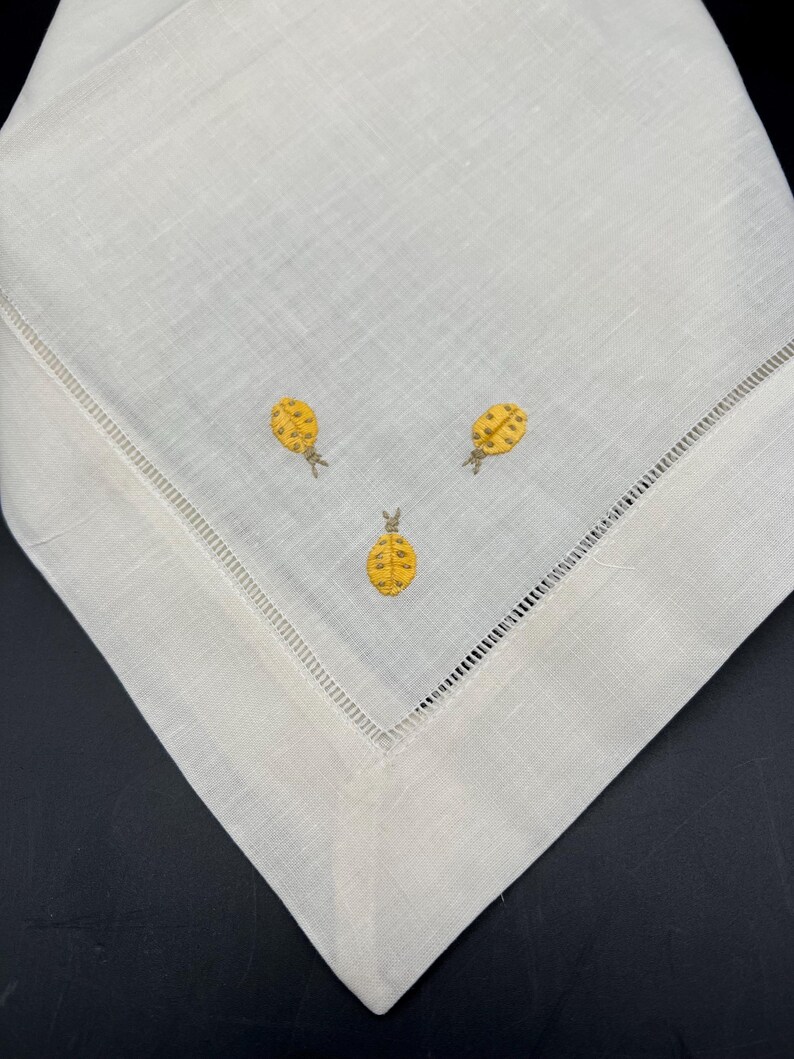 May include: A white linen napkin with a delicate embroidered design of three yellow pineapples.