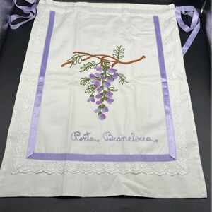 May include: White apron with a purple embroidered wisteria design and the text "Porto Branelva". The apron has a purple ribbon border and lace trim. The apron has purple ribbon ties.