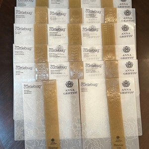 May include: A collection of rectangular Cricut Cuttlebug embossing folders by Anna Griffin. Each clear plastic folder features intricate embossed patterns, including floral, paisley, and geometric designs, with gold-toned branding strips.
