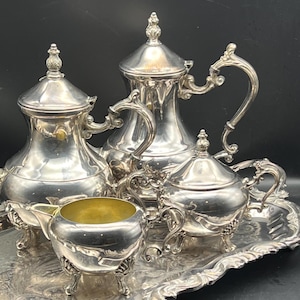 May include: A silver tea set with ornate detailing, including a teapot, creamer, sugar bowl, and serving tray. The set features curved handles and decorative accents. The interior of the creamer is gold-colored.