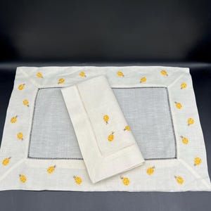 May include: A set of two white linen placemats with a yellow pineapple design embroidered around the border. The placemats are folded and stacked on top of each other.