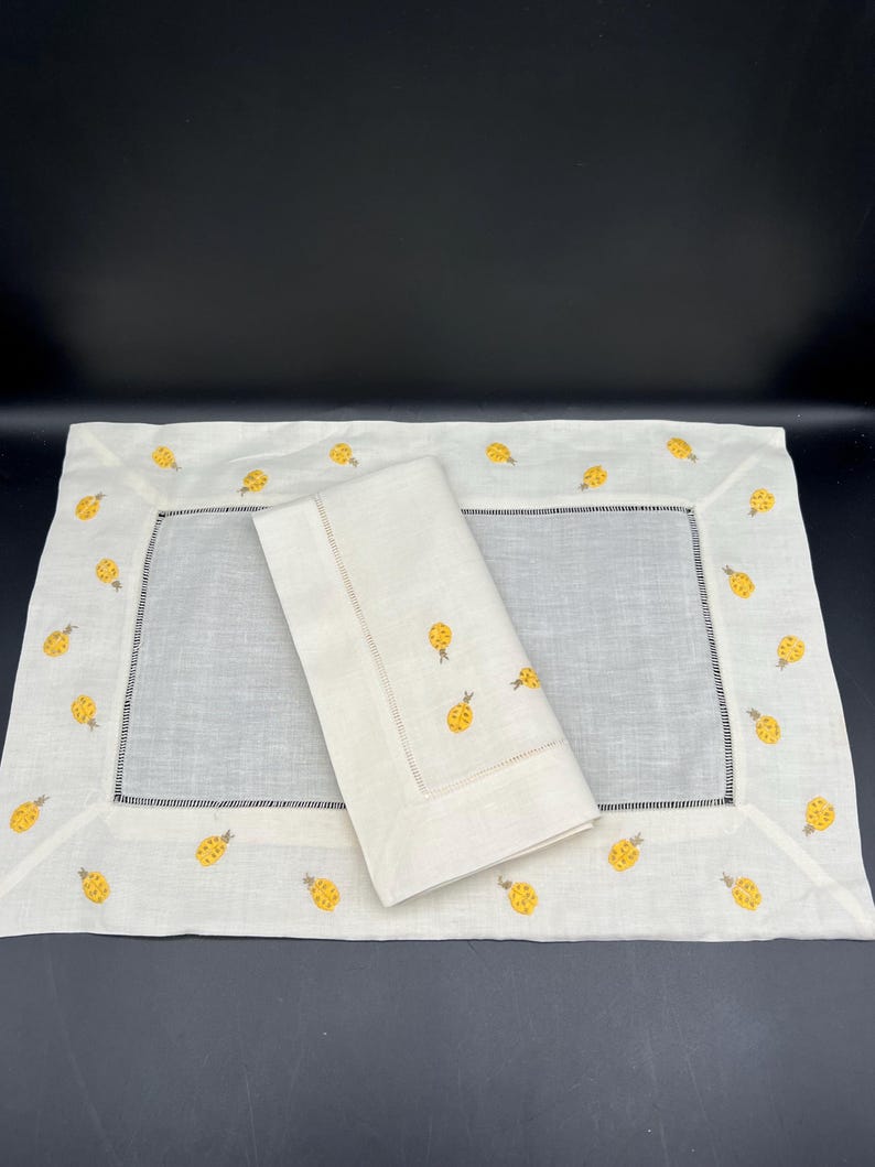 May include: A set of two white linen placemats with a yellow pineapple design embroidered around the border. The placemats are folded and stacked on top of each other.