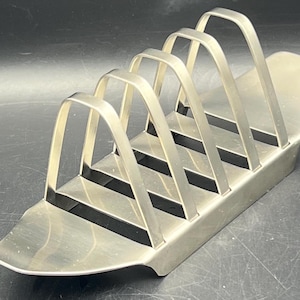 May include: A silver-colored metal toast rack with six arched dividers. The rack has a rectangular base and is designed to hold multiple slices of toast. The design is simple and functional, suitable for a kitchen or dining setting.