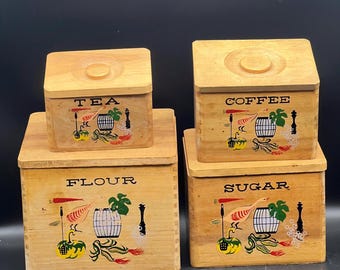 May include: Four vintage wooden kitchen canisters with lids. The canisters are labeled "TEA", "COFFEE", "FLOUR", and "SUGAR" in black block letters. Each canister features a painted design with a bird, barrel, and vegetables.