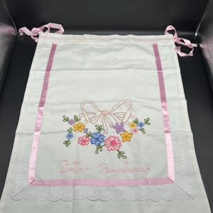 May include: White fabric bag with pink ribbon trim and embroidered floral design. Features a pink bow and colorful flowers. The text "Torta Biancheria" is embroidered below the bow. Pink ribbons are attached at the top.