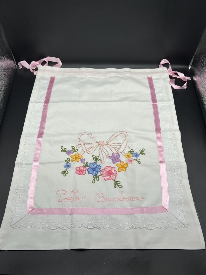 May include: White fabric bag with pink ribbon trim and embroidered floral design. Features a pink bow and colorful flowers. The text "Torta Biancheria" is embroidered below the bow. Pink ribbons are attached at the top.