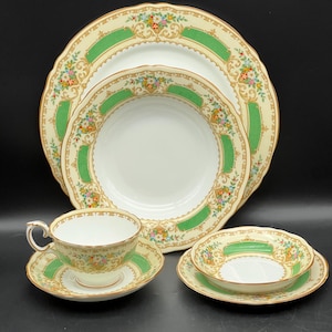 May include: A vintage cream-colored china set with a green and gold border. The set includes dinner plates, bowls, a teacup and saucer, and a small bowl. Floral accents decorate the edges of each piece. The set is arranged on a black surface.