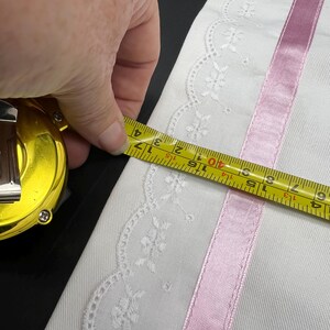 May include: White fabric with a delicate floral lace edge and a pink satin ribbon stripe. A yellow tape measure shows the fabric's width, measuring approximately 3 inches.