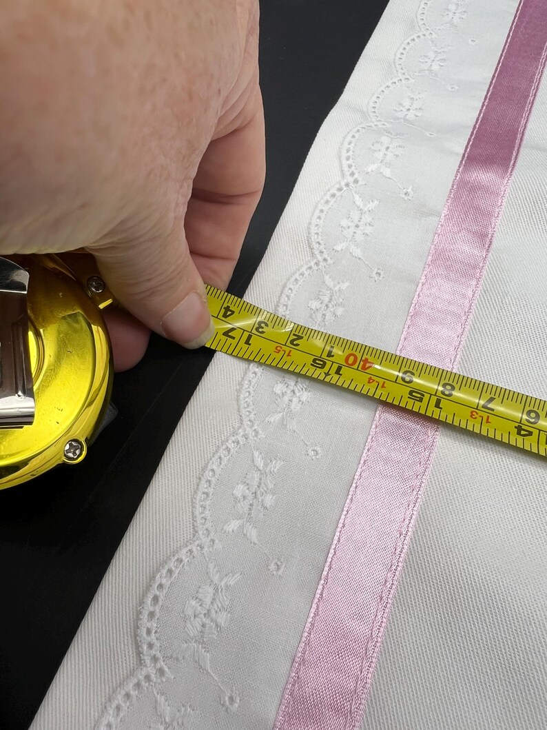 May include: White fabric with a delicate floral lace edge and a pink satin ribbon stripe. A yellow tape measure shows the fabric's width, measuring approximately 3 inches.