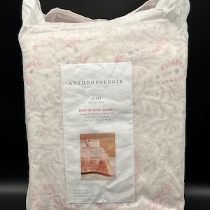 May include: A package of Anthropologie Josee Collection Pair of King Shams. The packaging is white with a floral pattern in pink and red. The label reads "Pair of King Shams" and shows a picture of the shams. The shams measure 20 x 36 inches.