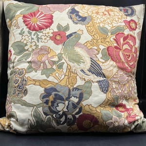 May include: Square decorative pillow with a floral and bird pattern. The pillow features a cream background with large pink, blue, and yellow flowers, and a green and blue bird. The design is reminiscent of vintage botanical prints.