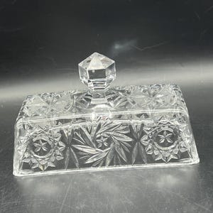 May include: A clear, rectangular glass butter dish with a faceted, diamond-shaped handle. The dish features a detailed, cut-glass pattern of stars and geometric shapes, adding a touch of elegance. The lid sits atop the base, showcasing the intricate design.