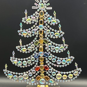 May include: A decorative tabletop Christmas tree sculpture constructed from a wire frame adorned with numerous iridescent, clear, and amber-colored faceted glass crystals and rhinestones, mounted on a rectangular metallic base.