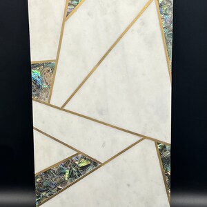 Op de afbeelding: Rechthoekig decoratief stuk van wit marmer met ingelegde abalone schelp accenten en goudkleurige metalen lijnen. Het ontwerp heeft geometrische vormen en patronen. De abalone schelp heeft iriserende kleuren. Het stuk staat op een zwarte achtergrond.