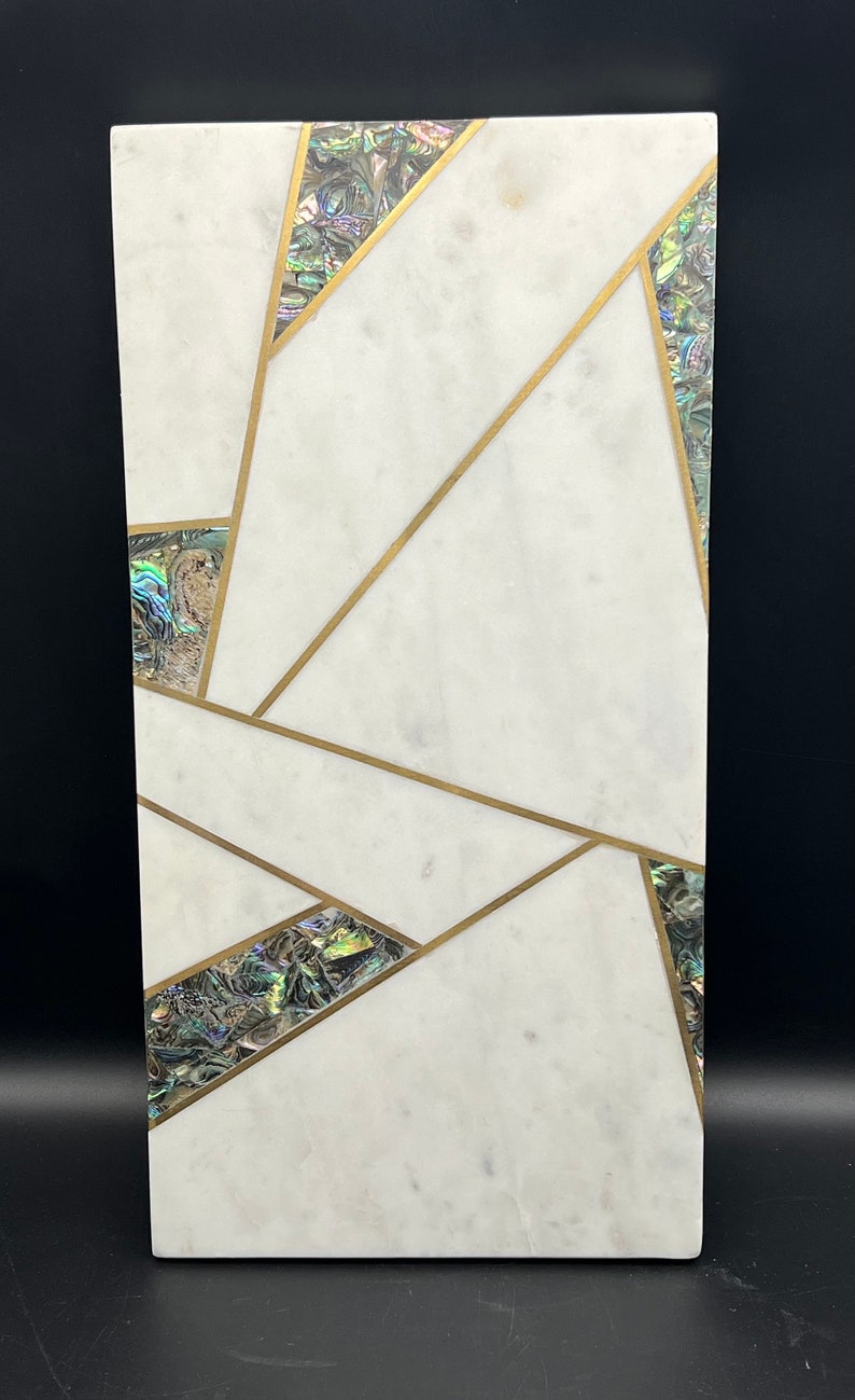 Op de afbeelding: Rechthoekig decoratief stuk van wit marmer met ingelegde abalone schelp accenten en goudkleurige metalen lijnen. Het ontwerp heeft geometrische vormen en patronen. De abalone schelp heeft iriserende kleuren. Het stuk staat op een zwarte achtergrond.