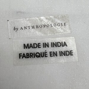 Op de afbeelding: Twee transparante labels met zwarte tekst. Het bovenste label luidt "by ANTHROPOLOGIE". Het onderste label vermeldt "MADE IN INDIA FABRIQU&Eacute; EN INDE". De labels liggen op een getextureerd, lichtgrijs oppervlak.