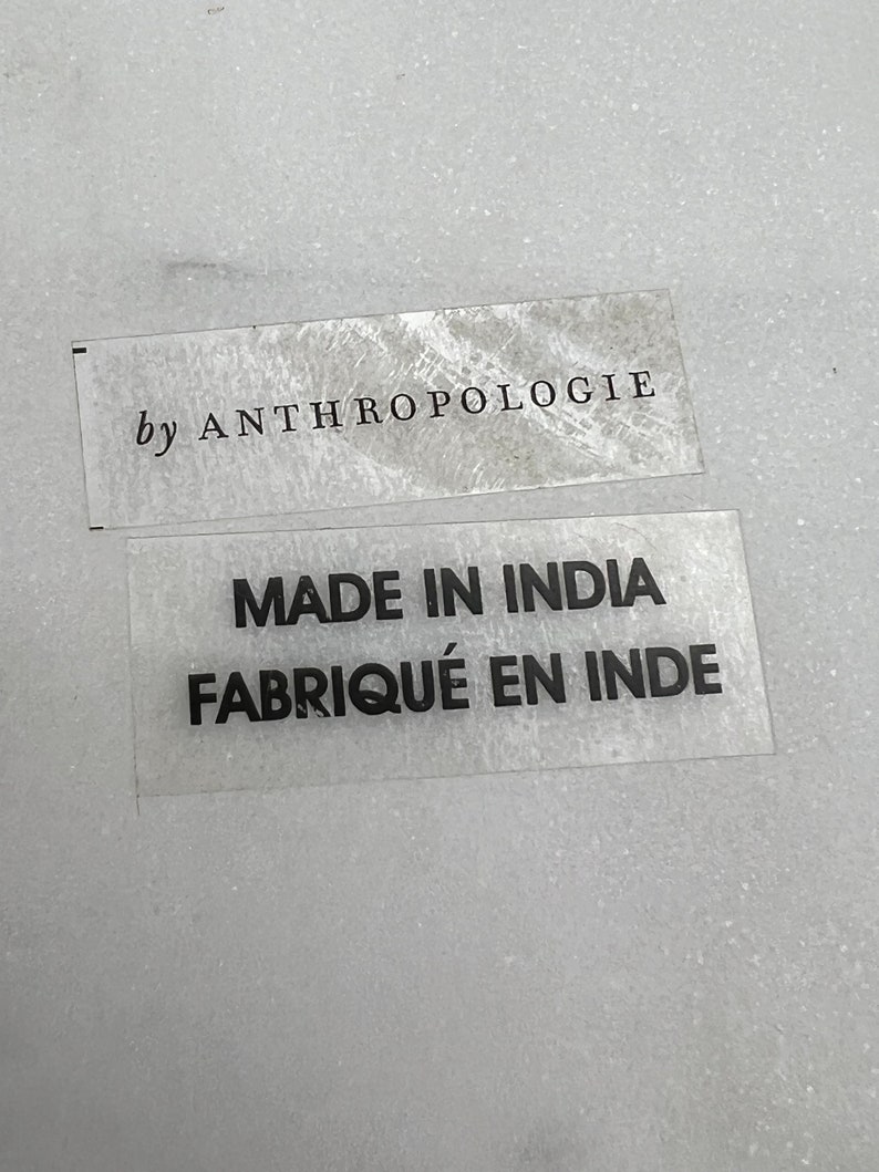 Op de afbeelding: Twee transparante labels met zwarte tekst. Het bovenste label luidt "by ANTHROPOLOGIE". Het onderste label vermeldt "MADE IN INDIA FABRIQU&Eacute; EN INDE". De labels liggen op een getextureerd, lichtgrijs oppervlak.