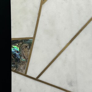 Op de afbeelding: Close-up van een wit marmeren oppervlak met ingelegd abalonschelp en messing accenten. Het ontwerp heeft geometrische vormen en lijnen, wat een moderne en elegante esthetiek cre&euml;ert. De abalonschelp voegt iriserende kleur toe.
