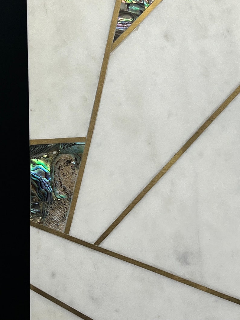 Op de afbeelding: Close-up van een wit marmeren oppervlak met ingelegd abalonschelp en messing accenten. Het ontwerp heeft geometrische vormen en lijnen, wat een moderne en elegante esthetiek cre&euml;ert. De abalonschelp voegt iriserende kleur toe.